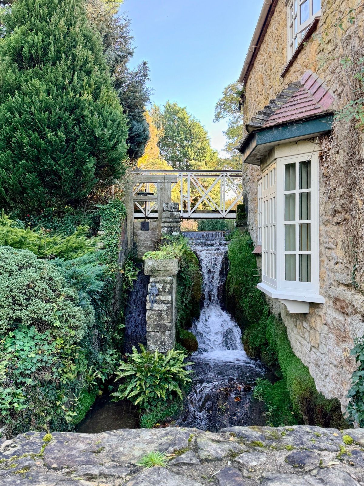 A small waterfall runs beside a stone cottage and wooden footbridge.